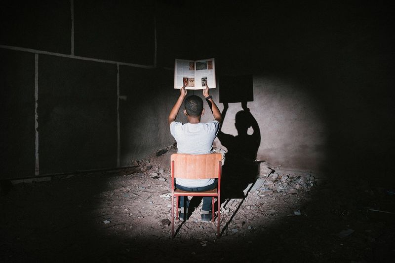 Una persona seduta su una sedia con le spalle rivolte verso l'obiettivo regge in alto un libro in una stanza buia, in una fotografia scattata con Canon EOS 5D Mark III da Mustafa Saeed.