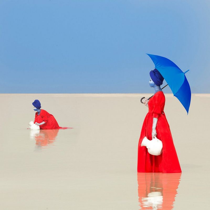 A woman in a red dress and holding a blue umbrella stands in shallow water holding a white jug. Another woman crouches behind her, filling her water jug.
