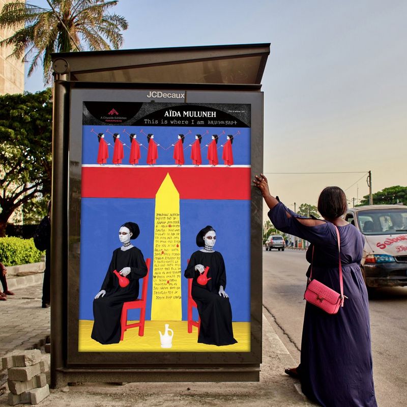 Una donna si appoggia a una fermata dell'autobus dove è esposta l'opera "This Is Where I Am" (2022) della fotografa Aïda Muluneh, mentre un'auto passa vicina.