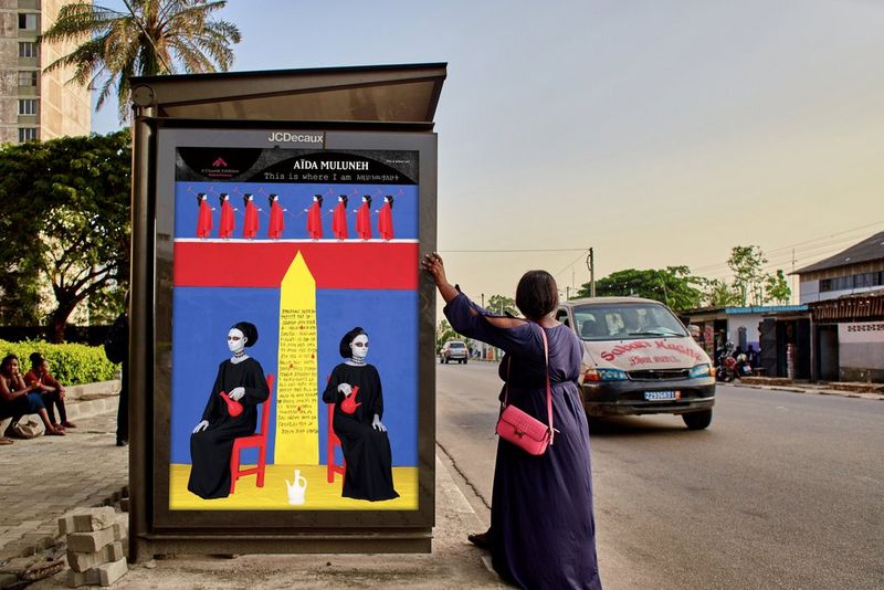 A woman leans up against a bus stop displaying photographer Aïda Muluneh's 2022 artwork This Is Where I Am as a car drives by.