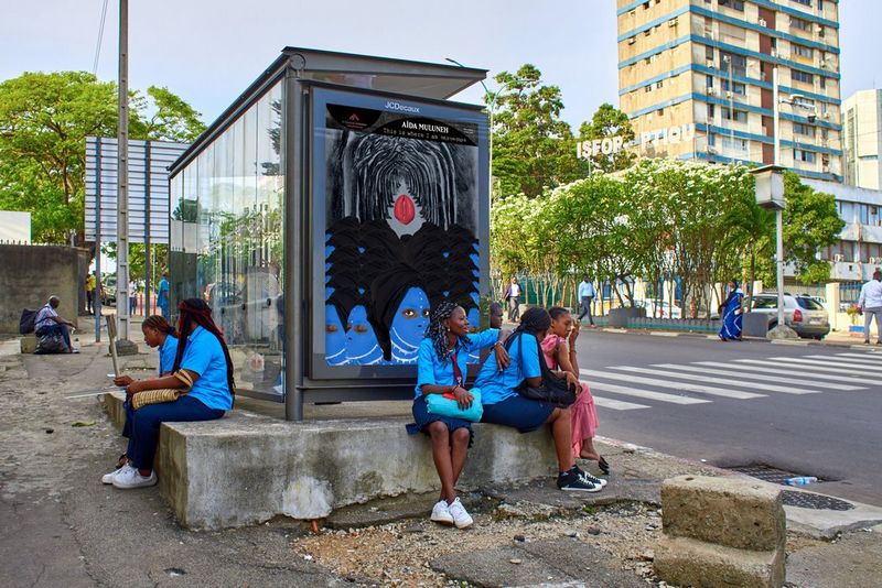 Figures sit on the pavement beside a bus shelter displaying Aïda Muluneh's 2022 artwork The Widows Of The Night, a road crossing and tall building in the background. 