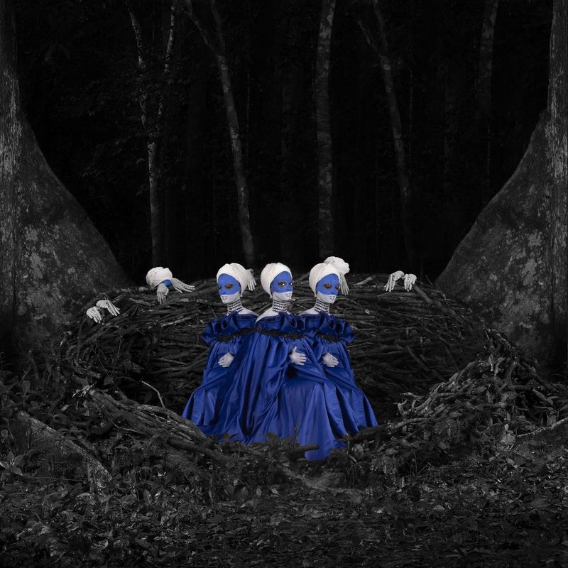 Trois femmes au visage peint en bleu, vêtues de robes bleues à volants, se tiennent debout dans une structure sombre ressemblant à un nid.