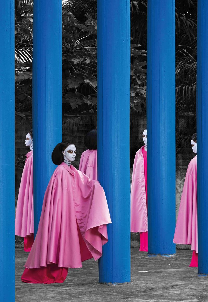 Représentations multiples de la même femme, vêtue d'une cape rose flottante et le visage peint en blanc, debout au milieu d'une série de colonnes d'un bleu vif.