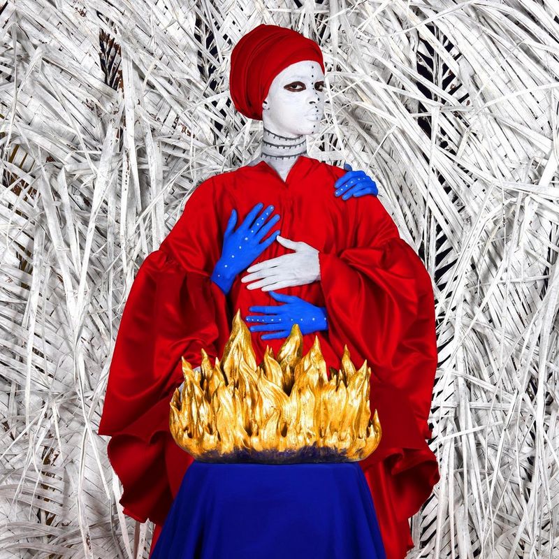 Une femme en tenue vietnamienne avec le visage peint en blanc se tient derrière une maquette représentant du feu, devant un décor de plantes et de feuilles blanches.