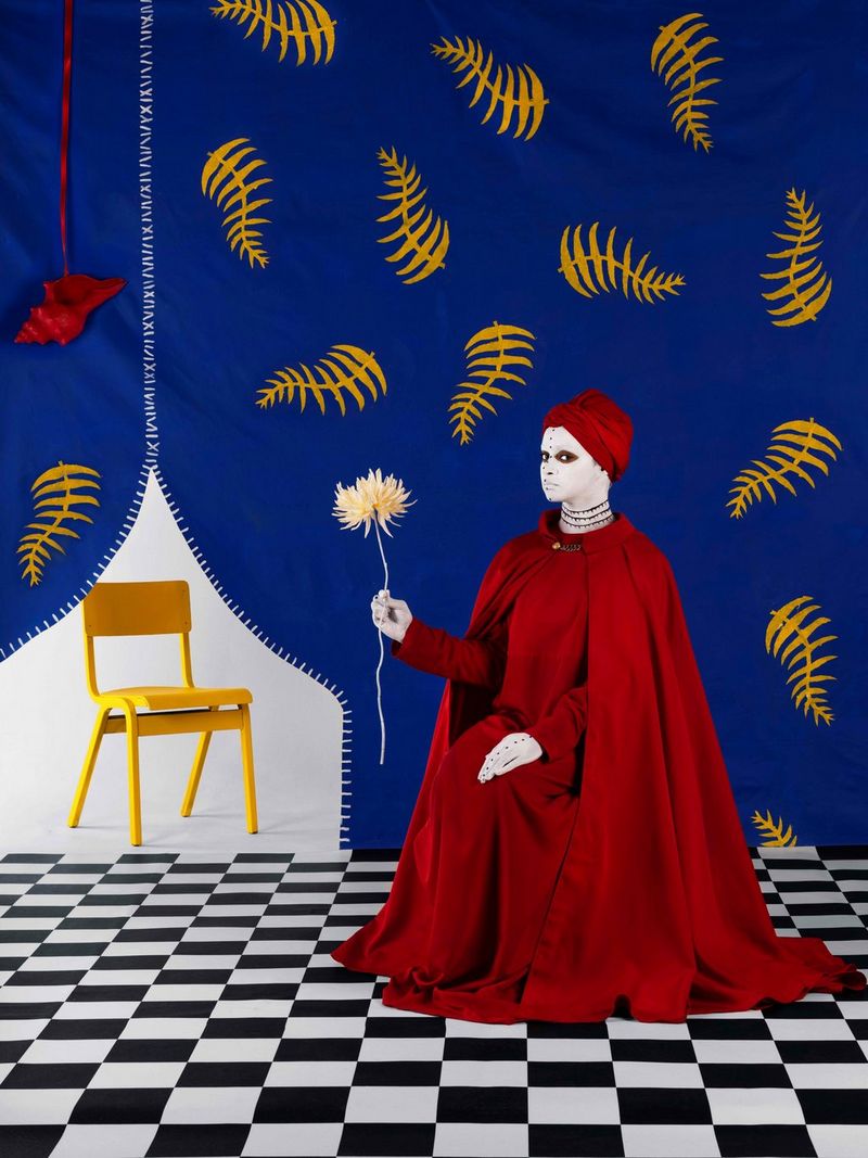 Une femme assise, vêtue d'une longue robe rouge foncé assortie d'une cape et d'un turban, se tient devant un fond bleu à motifs de feuilles et tient la tige d'une fleur.