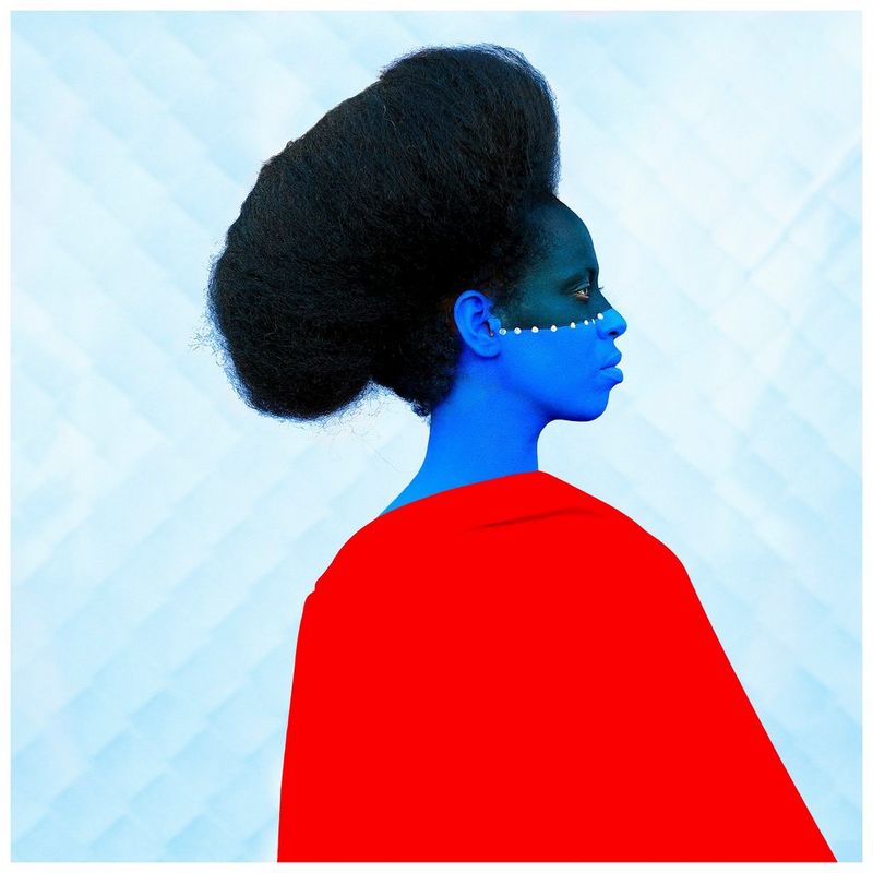 Une femme, coiffée d'une imposante coupe afro, se tient de profil avec le visage peint en bleu et une robe rouge.