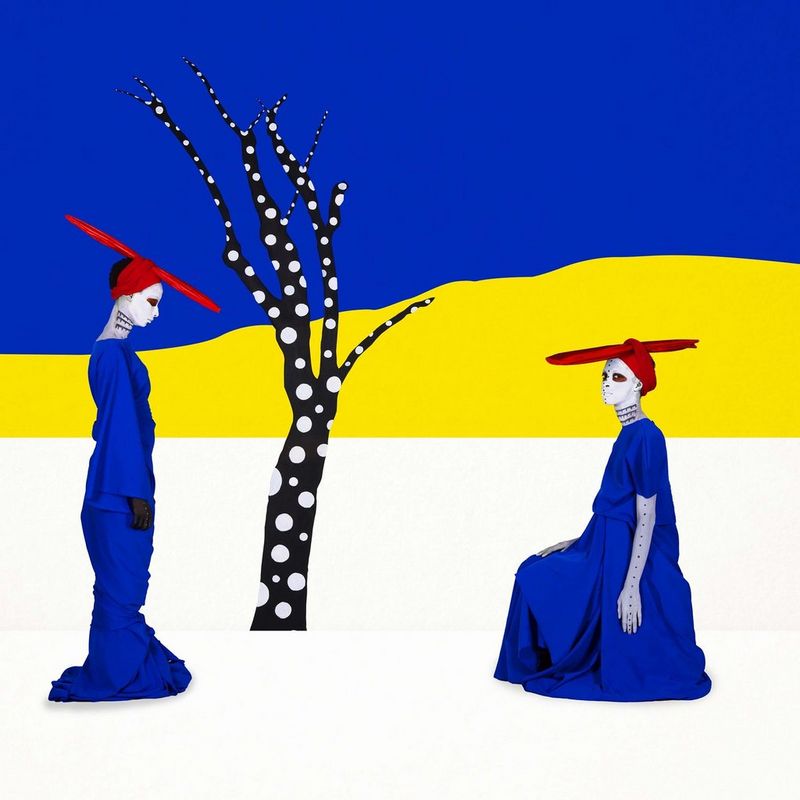 Two figures are seen in front of a stylised tree, both wearing a blue outfit and red hat, one kneeling in front of the other in Aïda Muluneh's 2020 artwork In Which We Remain.