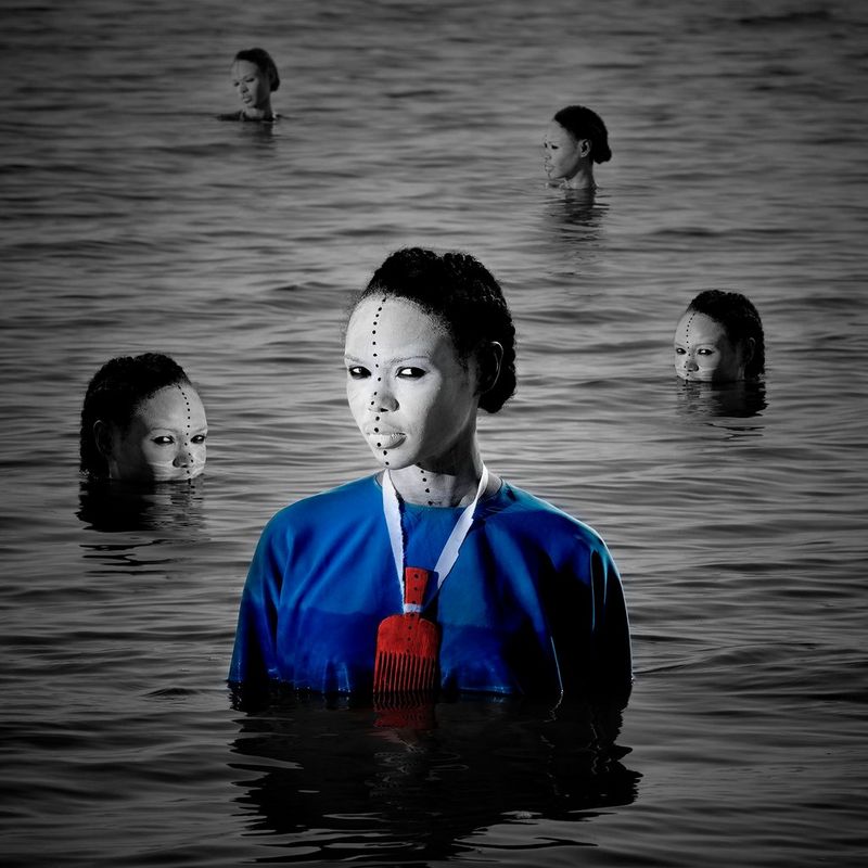 A woman in a blue outfit and white faceprint emerges from the water, other faces emerging behind her, in Aïda Muluneh's 2017 artwork The Return Of The Departure.