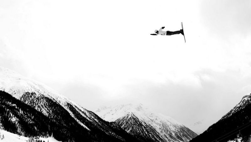 A black and white image of a skier flying through the air above snow-covered mountains.