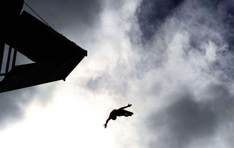 A black and white image of a cliff diver in action taken from below. The diver is silhouetted against a cloudy sky. 