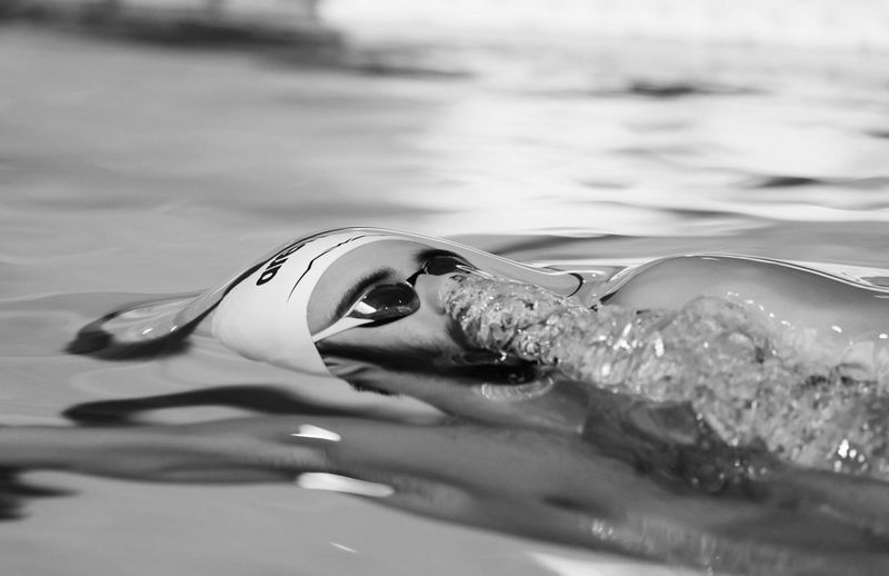A black and white image of a male swimmer's head emerging from the water, with a stream of bubbles emerging from his nose.