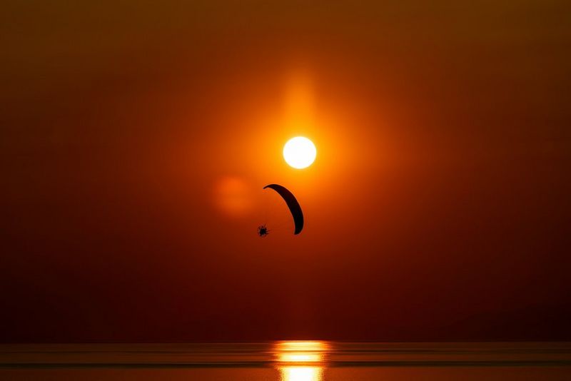 A paramotor pilot in silhouette against a red sky and a setting sun, the ocean below with the reflection of the sun on the water.