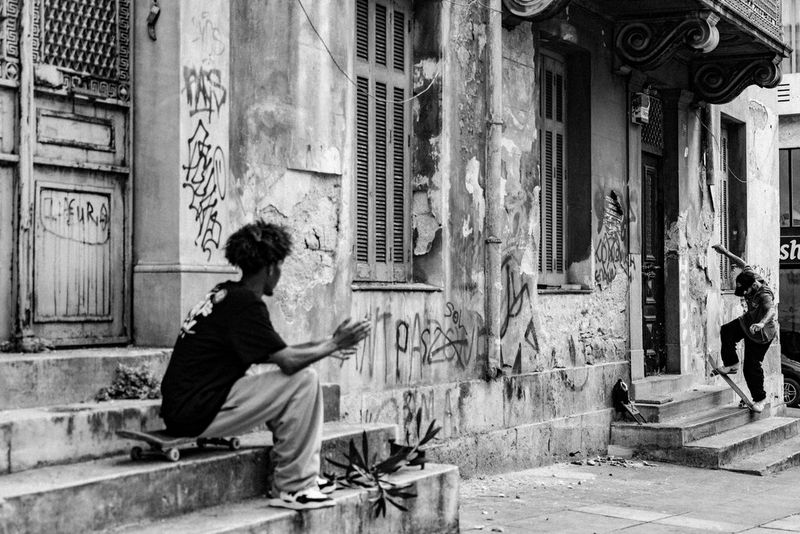 A black and white photo of two skateboarders in a street; one is performing a trick on the steps of a dilapidated building covered in graffiti, while the other looks on, clapping his hands.