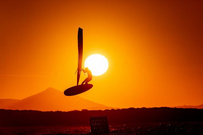 A windsurfer performs a stunt over a body of water, photographed against the sun so only his silhouette is visible. Mountains are visible in the background in a photo taken by Canon Ambassador Alexandros Grymanis. 