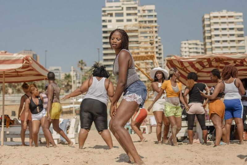 A woman dancing on a beach in Lebanon with a group of people under large, striped umbrellas behind her. 