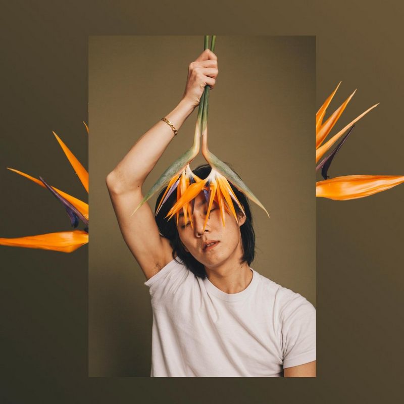 A man in a white t-shirt holds orange flowers over his face in front of a brown backdrop. The image is superimposed over another shot of the same flowers.