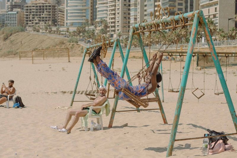 A male sunbather in a baseball cap watches a woman on a swing at a beach playground. 