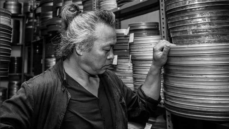 A black and white photograph of South Korean film director Kim Ki-duk, standing by shelves full of old film canisters. Taken on a Canon EOS 5D Mark II by Canon Ambassador Burak Günaydın.