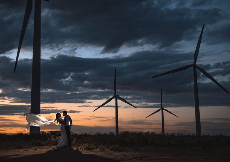 Surrounded by wind turbines, a couple embraces under a cloudy sky at sunset, the woman's white dress flowing in the wind. Taken on a Canon EOS 5D Mark IV by Canon Ambassador Askar Bumaga.