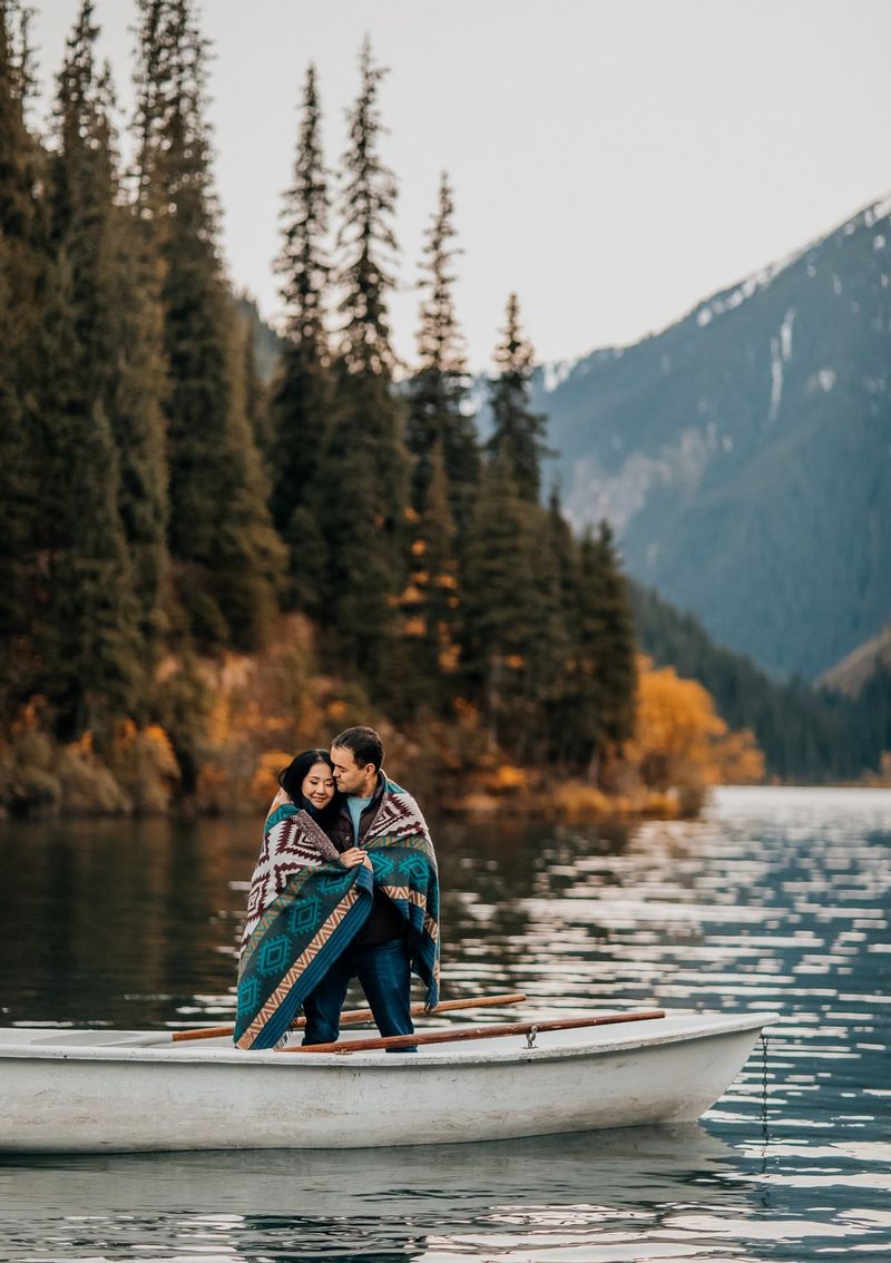 A couple embraces, wrapped in a blanket while standing on a boat, tall trees and a mountain visible in the background. Taken on a Canon EOS 5D Mark IV by Canon Ambassador Askar Bumaga.