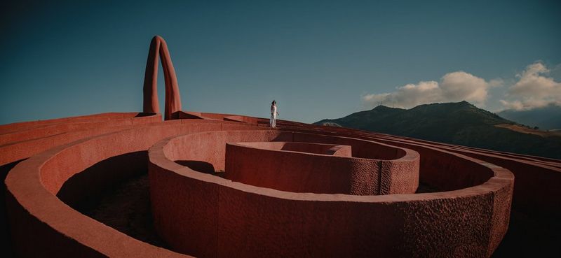 A woman in a long white dress stands on top of a vast sculptural labyrinth that twists and curves in on itself, with a large mountain range in the background.