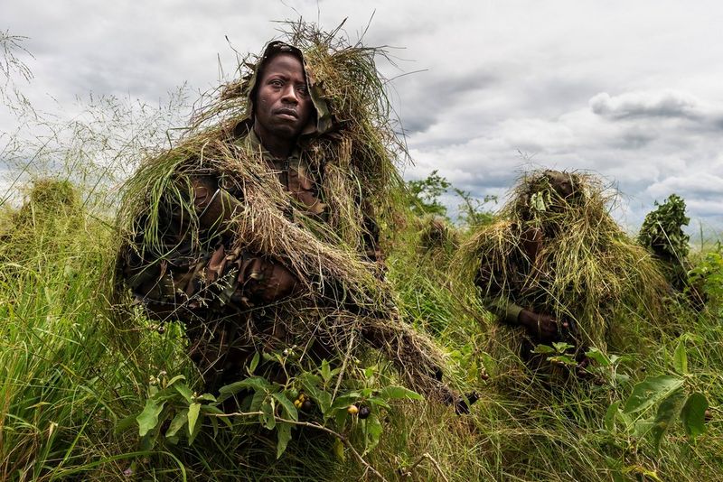 Armed conservation rangers emerge from the bush wearing uniforms camouflaged with grass and foliage.