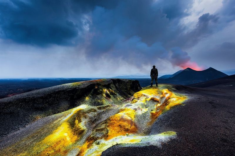 A silhouetted man stands on top of a new lava field that glows bright yellow. Smoke billows from a volcano in the background.