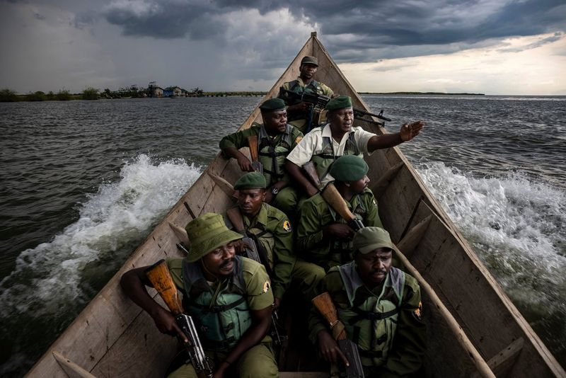 A group of men in green uniforms and holding guns sit in the prow of a boat.