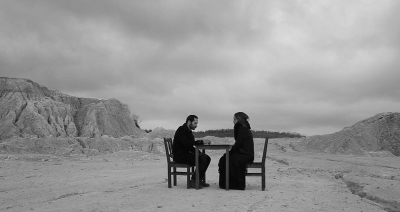 A black and white still from Canon Ambassador Çağla Çağlar's first short film Minval, shot on a Canon EOS 7D, showing two people sitting at a table in a desert landscape.