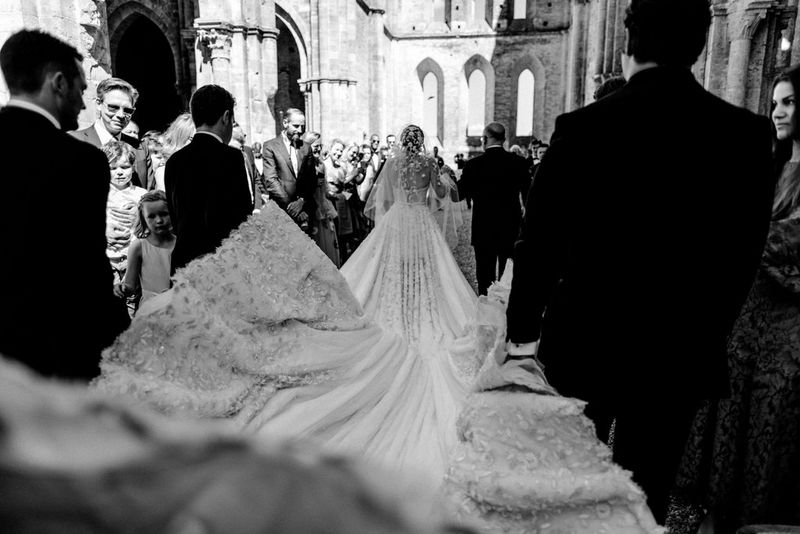 A black and white image, shot from behind, of a bride walking down the aisle of a church in a dress with a very long train.