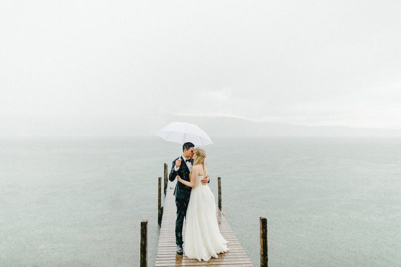 A bride and groom kiss under an umbrella on a small pier at the edge of a lake. 