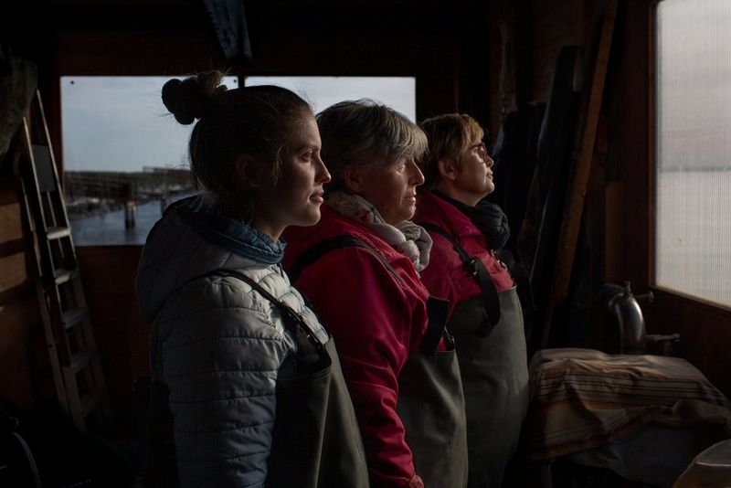 Side profile of three fisherwomen, of varying ages, standing side by side in a small boathouse. All three are wearing warm, waterproof clothing and smiling slightly. Taken on a Canon EOS 5D Mark III by Chiara Negrello.