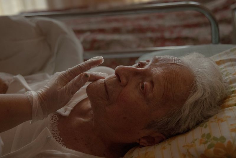 A hand wearing a clear plastic glove strokes the face of an elderly woman in a hospital bed. Taken on a Canon EOS R5 by Chiara Negrello.