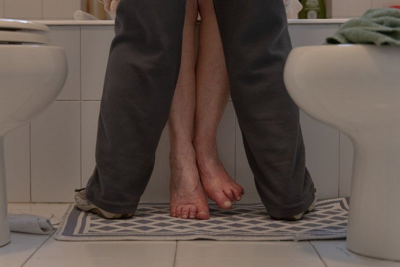 Framed from the knees down, a caregiver wearing grey jogging bottoms helps an elderly woman into a bathtub. Taken on a Canon EOS R5 by Chiara Negrello.