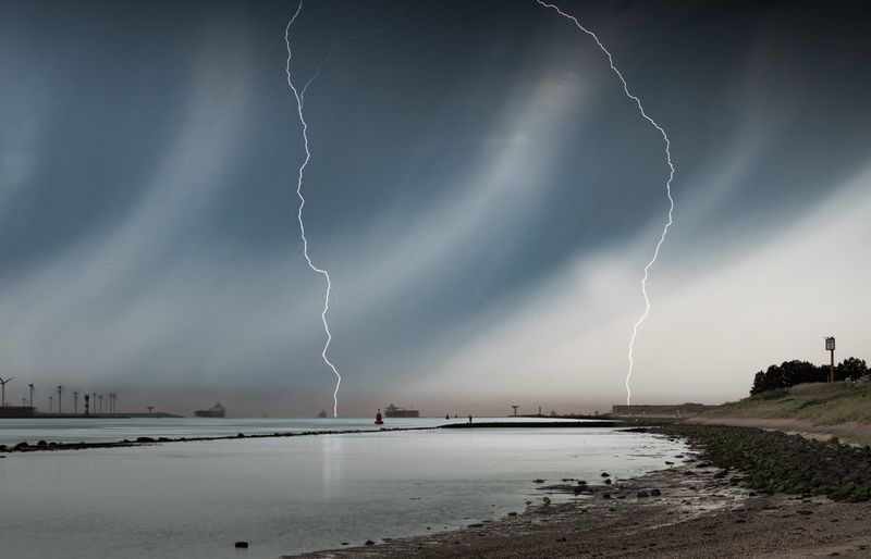 Two lightning strikes descend from a dark storm front over calm coastal waters.