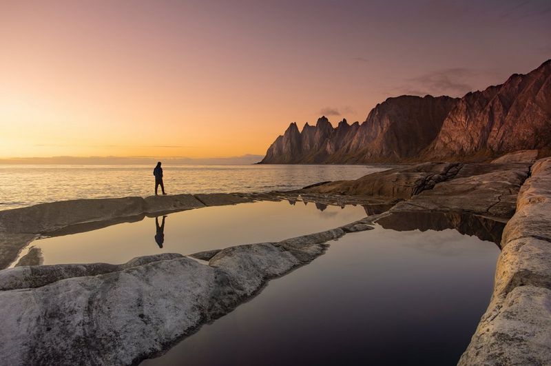 A lone figure standing within a reflective coastal landscape at sunset.