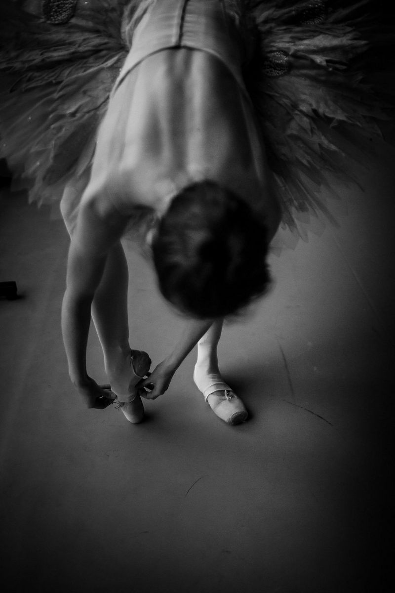 A black and white portrait of a ballerina bending down to pull her tights over her ballet shoe.