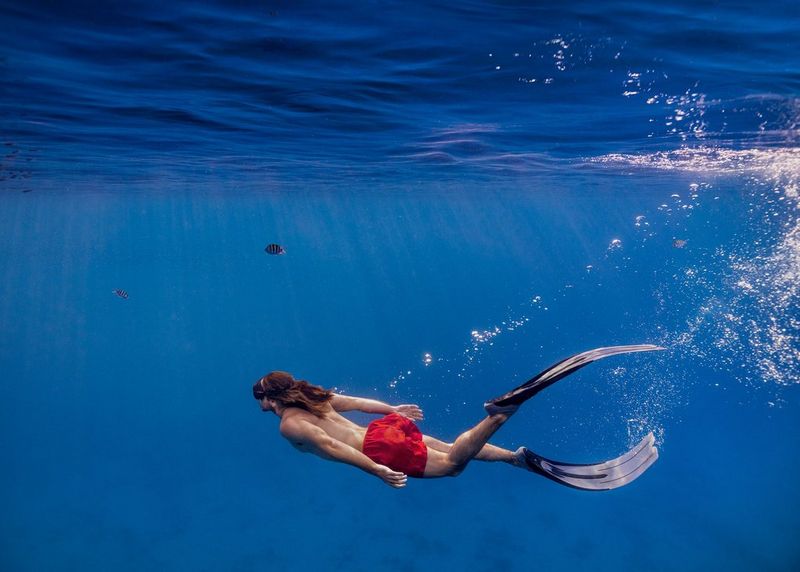 A young man with long hair freediving in the Red Sea while wearing red trunks and flippers.
