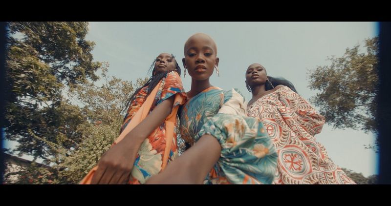 A still, shot from below, from footage captured by filmmaker Daniel Ehimen on a Canon EOS C70 with a Canon CN-E14mm T3.1 L F lens, showing three women in colourful dresses looking down at the camera.