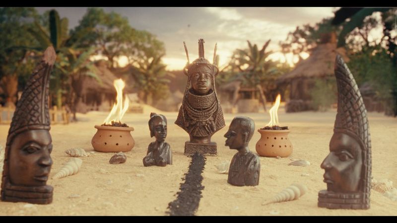 A still from footage shot by filmmaker Daniel Ehimen showing African sculptures arranged on a sandy surface alongside shells and two fire bowls.