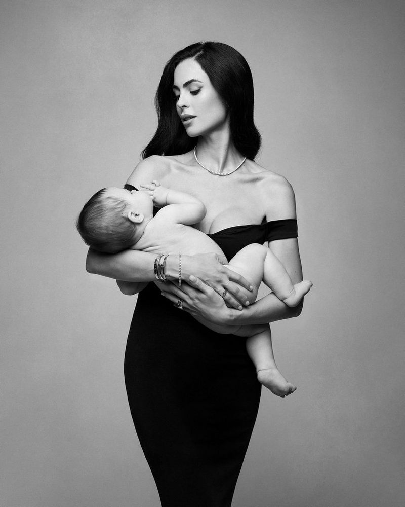 A black and white portrait of a woman in a black off-the-shoulder dress gazing lovingly at the naked baby she is holding, taken on a Canon EOS R5 with a Canon RF 28-70mm F2L USM lens by Donatella Nicolini.