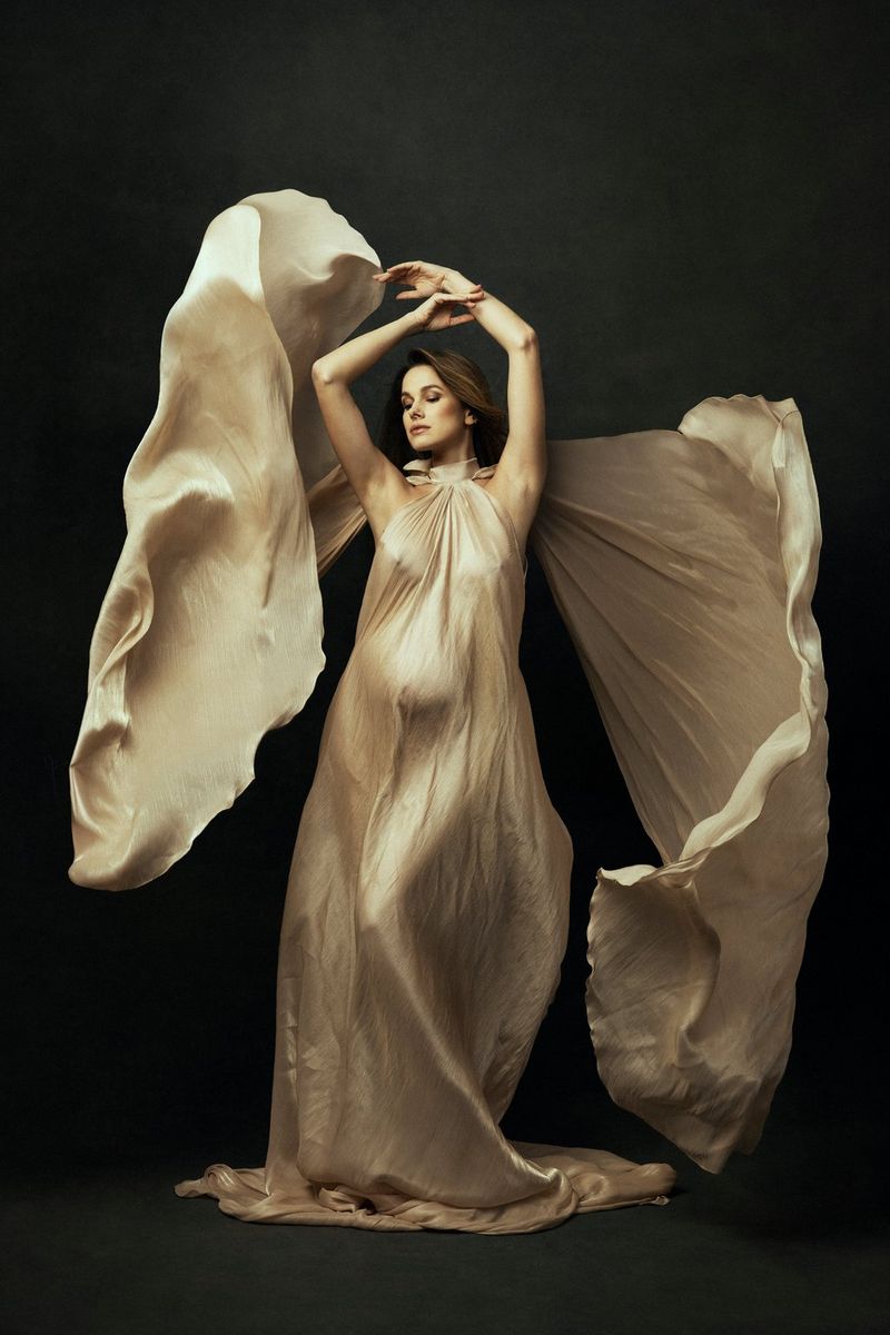 A portrait of a pregnant woman with her arms raised above her head and the train of her cream satin dress arranged wing-like behind her.