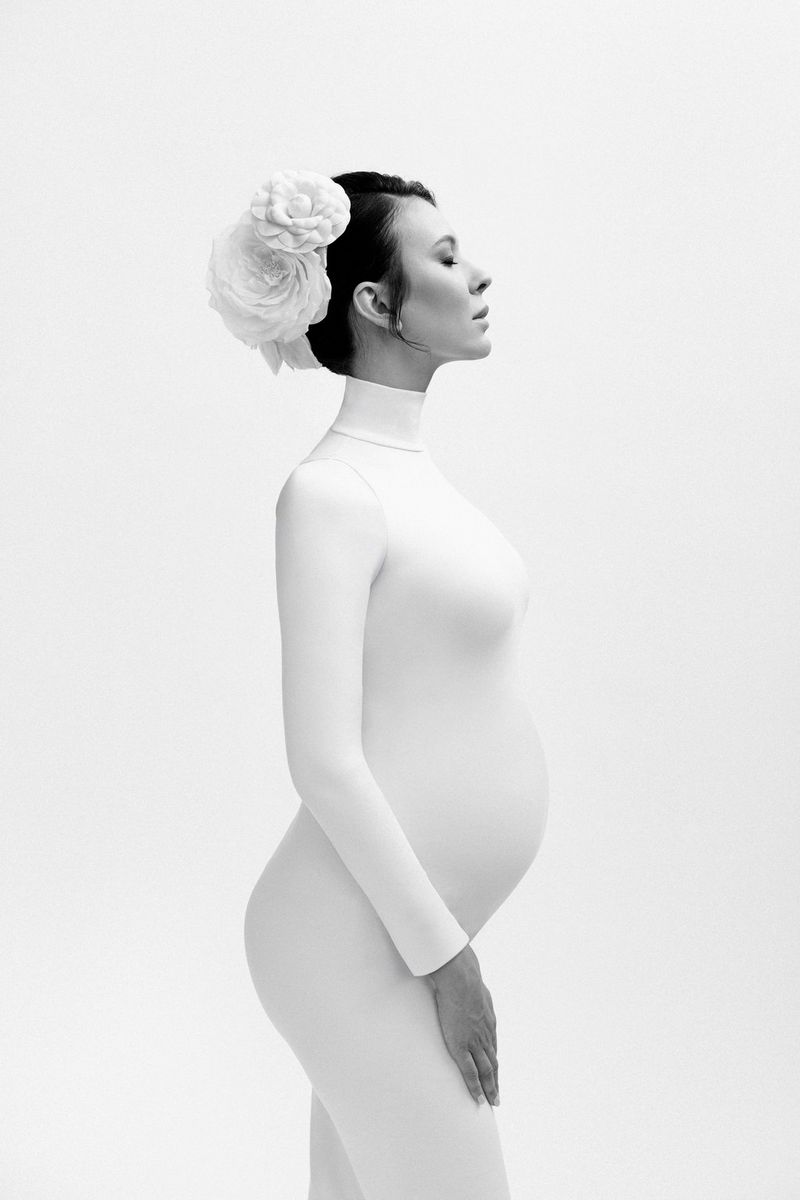 A black and white portrait of a pregnant woman in side profile wearing a white high-neck dress and with flowers in her hair.