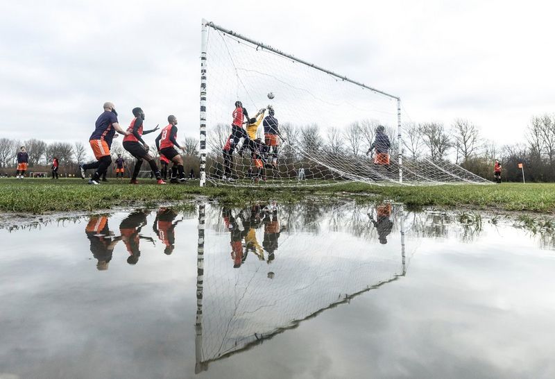 Men playing football on a muddy pitch, the players reflected in a large puddle of water behind the goal.