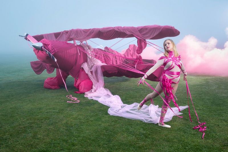 A woman wearing a beige leotard and ballet shoes with pink ribbons tied around her legs, hands and upper body stands next to a small plane also draped in bright pink.