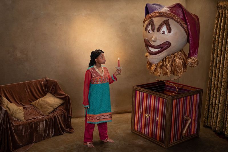 A young girl holding a candle looks defiantly at an oversized jack-in-the-box children's toy.