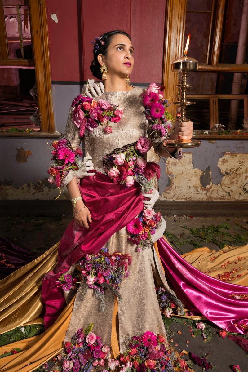 A woman wearing a long dress covered in flowers and with several trains holds up a lit candelabra. She is looking away from the camera and several white hands can be seen grabbing her body.