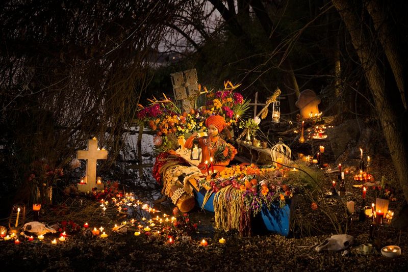 A young woman sits in a boat covered in flowers, drums and animal skins. She is holding a lantern and in the water and on the bank around her are hundreds of lit candles.