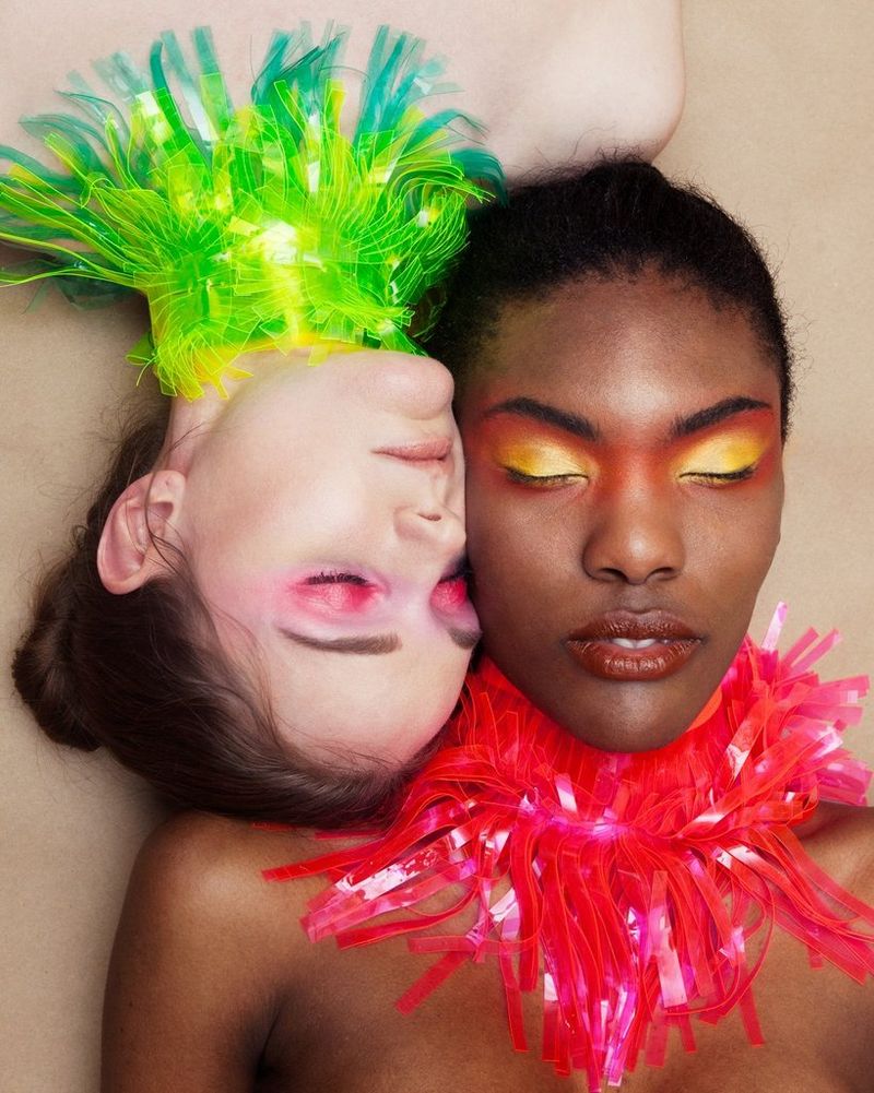  Two models wearing colourful eyeshadow and elaborate neck pieces lie down with their heads together and their eyes closed.
