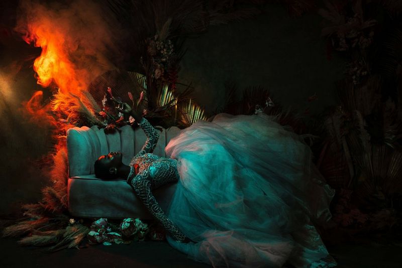 Une mannequin vêtue d'une robe de tulle avec un corsage à paillettes est allongée sur un sofa. Des flammes semblent lécher le mur derrière elle.